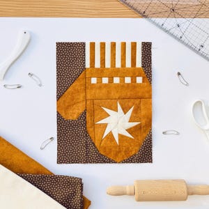 Peut inclure: Un motif de mitaine en patchwork marron et beige, orné d'une étoile. L'image comprend des outils de couture tels que des ciseaux, des épingles, une règle et un rouleau à pâtisserie, suggérant un projet de quilting ou de couture. Des échantillons de tissu sont également présents.
