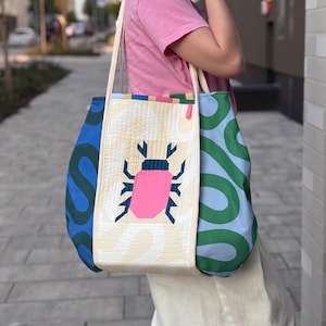 May include: A quilted tote bag featuring a central cream panel with a pink and dark blue beetle graphic. The side panels are light blue and dark blue with a green wavy pattern. The bag has two long shoulder straps and a rectangular construction.