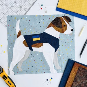 May include: A blue and white fabric patchwork quilt block depicting a dog with a blue and yellow patch on its chest. The dog is surrounded by a blue background with white stars.