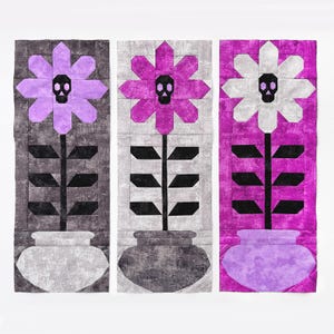 May include: Three rectangular fabric quilt blocks featuring potted flowers with skull centres. The left block has a light purple flower in a grey pot, the centre has a magenta flower in a dark grey pot, and the right has a white flower in a purple pot.