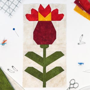 May include: A handmade quilt block featuring a stylized red flower with a yellow center and green stem and leaves. The flower is made of geometric shapes and set against a neutral background. Sewing notions are scattered around the block.