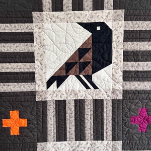 May include: A quilt block with a black and brown bird design, a pink and purple flower design, and a white and orange flower design. The quilt block is made of fabric with a white and gray striped pattern.