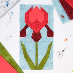 May include: A fabric block featuring a geometric tulip design. The tulip has a red patterned head and dark red stem and petals, set against a light blue background. Green leaves extend from the stem. Sewing notions and fabric scraps are scattered around the block.