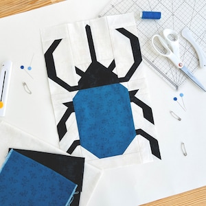 May include: A white fabric block with a black and blue geometric design of a beetle. The fabric block is on a white surface with sewing tools and fabric scraps.