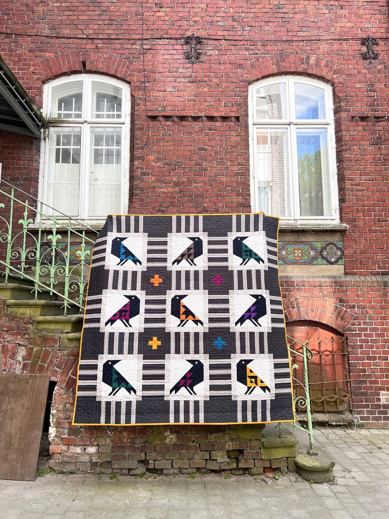 May include: A black and white quilt with a geometric pattern of birds. The birds are made of different colored fabrics, including red, orange, yellow, green, blue, and purple. The quilt is hanging on a brick wall.