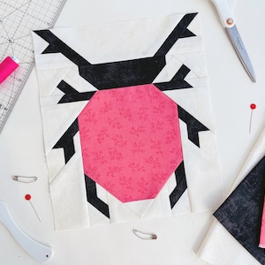 Puede incluir: Un bloque de colcha de retazos de tela rosa y negro con un diseño geométrico que se asemeja a un escarabajo. El bloque está hecho de diferentes telas con un fondo blanco y acentos rosa y negro.