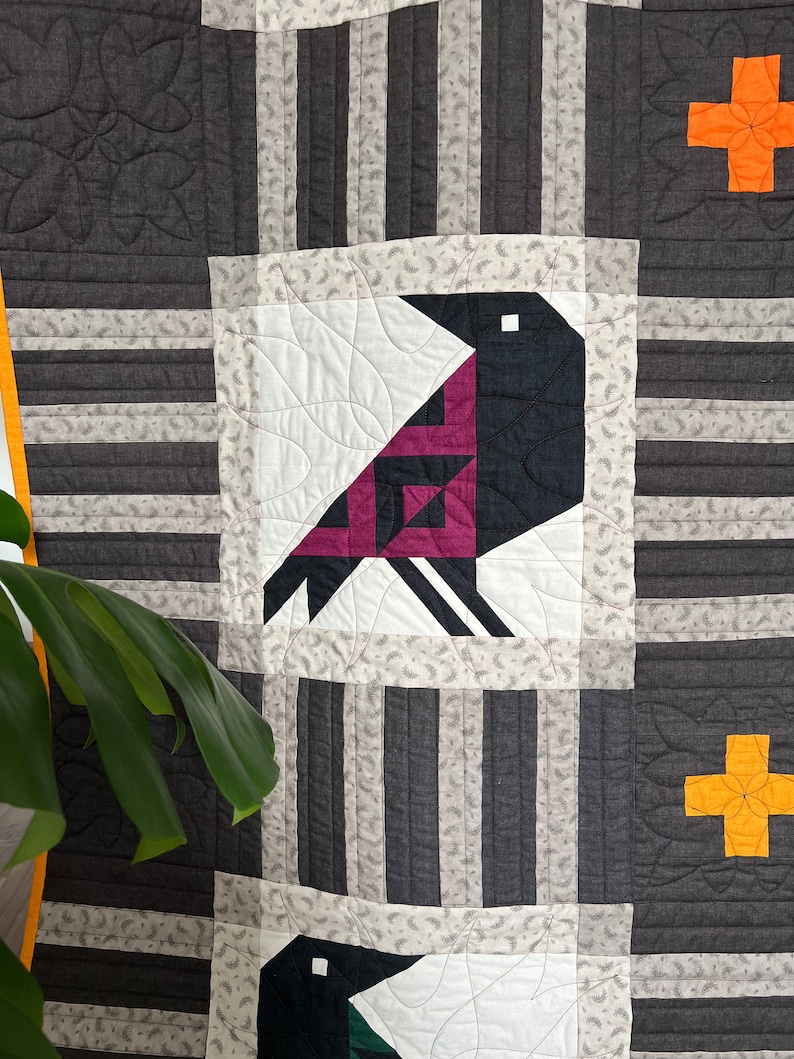 May include: A quilt with a geometric pattern featuring a black and pink bird, a yellow plus sign, and gray and white stripes.