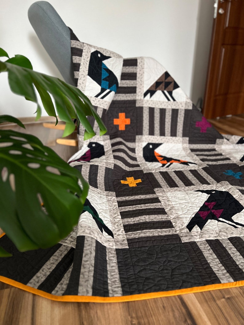 May include: A black and white quilt with a geometric pattern of birds and crosses. The quilt is made with a variety of fabrics, including a black and white striped fabric, a solid black fabric, and a solid white fabric. The quilt is edged with a bright orange trim.