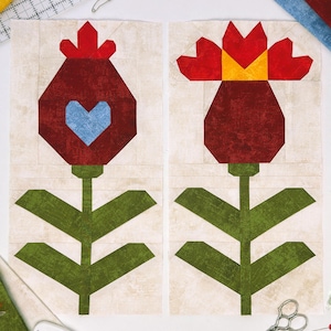 May include: Two fabric flower blocks. One features a red flower with a blue heart, the other a red flower with a yellow center. Both have green stems and leaves. The background is a light beige.