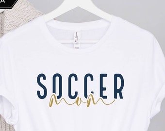 Może przedstawiać: Biała koszulka z napisem "SOCCER MOM" w kolorze granatowym i złotym. Tekst "SOCCER" jest w pogrubionej, blokowej czcionce. Słowo "MOM" jest w czcionce kursywą. Koszulka wisi na białym wieszaku.