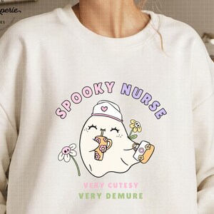 Puede incluir: Una sudadera blanca con un diseño gráfico de un fantasma con un gorro de enfermera y sosteniendo una taza y una pequeña bolsa. El texto "SPOOKY NURSE" está por encima del fantasma y el texto "VERY CUTESY VERY DEMURE" está por debajo del fantasma. El texto "PNG FOR SUBLIMATION" está en la parte inferior de la imagen.