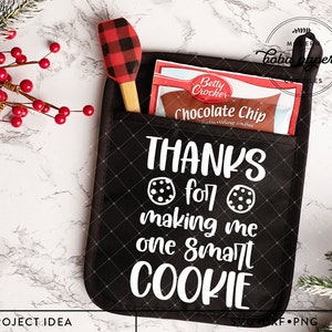 Pode incluir: Luva de forno preta com texto branco que diz "THANKS for making me one SMART COOKIE". Duas imagens de biscoitos com gotas de chocolate estão de cada lado do texto. Uma espátula vermelha e preta xadrez e um pacote de mistura para biscoitos com gotas de chocolate estão no bolso da luva de forno.