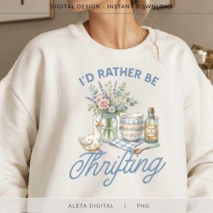 Puede incluir: Sudadera color crema con una ilustración en acuarela. El diseño presenta un ramo de flores en un tarro, un pato y cuencos. El texto dice "I'd Rather Be Thrifting" en escritura azul.