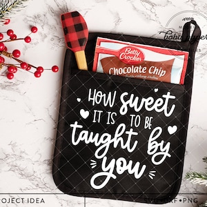 以下が含まれることがあります： 「How sweet it is to be taught by you」と白文字で書かれた黒いオーブンミット。ミットには、チョコレートチップクッキーミックスのパッケージと赤と黒のヘラが入ったポケットがあります。