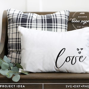 May include: White pillow with black cursive text that says "love" and three small black hearts above the word. The pillow is on a wooden bench with a black and white plaid pillow behind it.