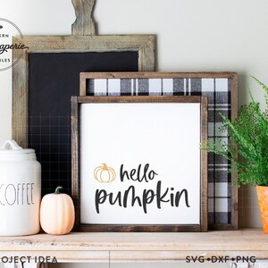 Op de afbeelding: Een wit bord met een zwarte houten lijst en de tekst "hello pumpkin" met een kleine oranje pompoenafbeelding boven de tekst. Het bord staat op een houten plank met een koffiepot, een kleine oranje pompoen en een plant in een pot.