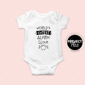 May include: White baby onesie with black text that reads "World's Cutest Alarm Clock" with a cartoon alarm clock graphic. The onesie has a Project Idea logo in the bottom right corner.