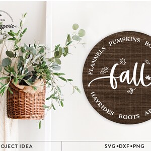 May include: A round wooden sign with the word "fall" in white script surrounded by a circle of fall-themed words like "flannels", "pumpkins", "bonfires", "hayrides", "boots", and "apple cider".