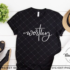 以下が含まれることがあります： 白い木製の表面に帽子と靴を置いた、黒いTシャツ。Tシャツには、白い筆記体で「worthy」という文字が書かれており、その上に十字架が描かれている。