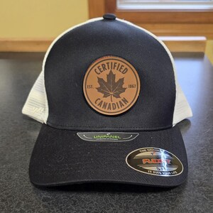 May include: A black and white baseball cap with a brown leather patch that says "Certified Canadian Est. 1867". The patch features a maple leaf.
