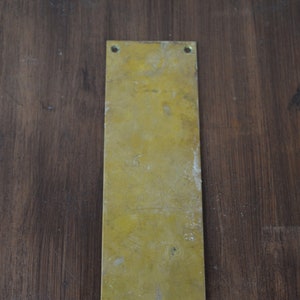 Può includere: Una placca per porta rettangolare in ottone con quattro fori per viti. La placca è leggermente usurata e ha una finitura patinata.