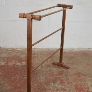 May include: A freestanding wooden towel rack featuring a medium brown finish. The structure consists of two vertical supports with T-shaped feet, connected by three horizontal bars arranged in a tiered formation for hanging items.
