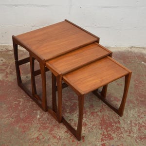 May include: A set of three nesting tables crafted from medium-toned wood. Each table features a rectangular top with slightly raised edges and four curved legs that connect to form a continuous frame on each side, allowing them to slide together.
