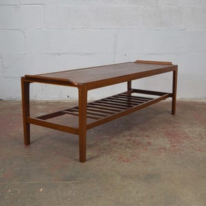 May include: A rectangular brown wooden coffee table featuring a smooth top surface with slightly raised edges on the short sides and a slatted lower shelf supported by four straight legs, showcasing a natural wood grain finish throughout.