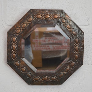 Pode incluir: Um espelho octogonal com uma moldura de metal escuro martelado, apresentando um padrão em relevo de formas ovais conectadas por linhas finas. A moldura tem um acabamento envelhecido com reflexos acobreados nos elementos decorativos.