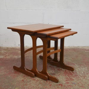 Può includere: Un set di tre tavolini a incastro realizzati in legno di tonalità media. Ogni tavolo presenta un piano rettangolare con bordi arrotondati e una base composta da due supporti verticali collegati da un traversino orizzontale curvo.