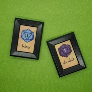 May include: Two framed cross-stitch pieces with blue and purple dice and the words "Whelp" and "Oh shit" on a green background.