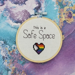 以下が含まれることがあります： 「This is a Safe Space」という文字と、中央に虹色のハートが描かれた白い刺繍枠です。