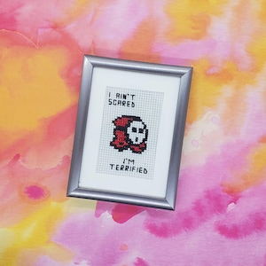 Pode incluir: Um ponto de cruz enquadrado com uma imagem pixelada de um personagem vermelho e preto com uma máscara de caveira. O texto diz "I ain't scared I'm terrified".