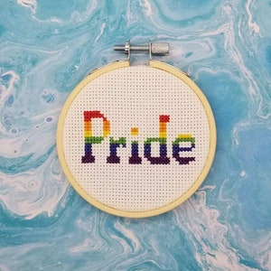 Può includere: Un cerchio da ricamo bianco con la parola "Pride" ricamata in colori arcobaleno.