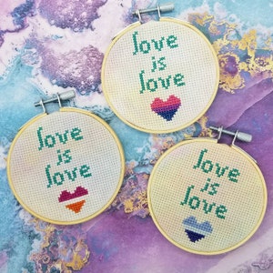 Op de afbeelding: Drie borduurringen met de woorden "love is love" in groene draad. Elke ring heeft een hart in verschillende kleuren, die verschillende LGBTQ+-vlaggen vertegenwoordigen. De ringen staan tegen een kleurrijke, gemarmerde achtergrond.