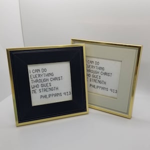 May include: Two framed cross-stitch artworks with gold trim. One frame is black, the other is off-white. Both feature the text "I CAN DO EVERYTHING THROUGH CHRIST WHO GIVES ME STRENGTH PHILIPPIANS 4:13" in black.