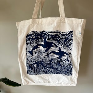 Puede incluir: Una bolsa de lona blanca con una ilustraci&oacute;n azul y blanca de dos orcas nadando en el oc&eacute;ano. La ilustraci&oacute;n incluye algas marinas, medusas y otras formas de vida marina. La bolsa tiene un asa larga y est&aacute; hecha de algod&oacute;n natural.