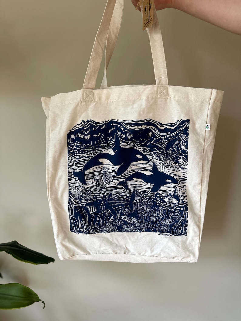 Puede incluir: Una bolsa de lona blanca con una ilustraci&oacute;n azul y blanca de dos orcas nadando en el oc&eacute;ano. La ilustraci&oacute;n incluye algas marinas, medusas y otras formas de vida marina. La bolsa tiene un asa larga y est&aacute; hecha de algod&oacute;n natural.