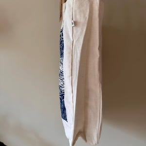 Puede incluir: Una bolsa de lona blanca con un estampado gr&aacute;fico azul y blanco. La bolsa tiene un asa de corcho y una peque&ntilde;a etiqueta verde.