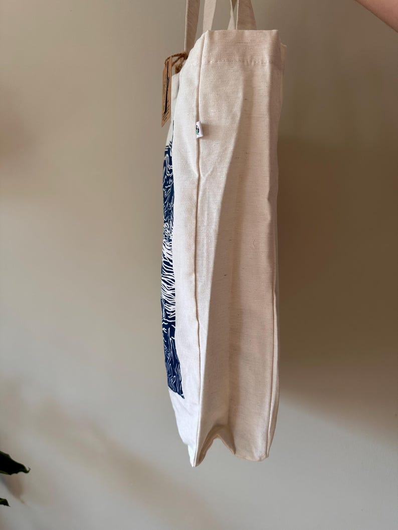 Puede incluir: Una bolsa de lona blanca con un estampado gr&aacute;fico azul y blanco. La bolsa tiene un asa de corcho y una peque&ntilde;a etiqueta verde.