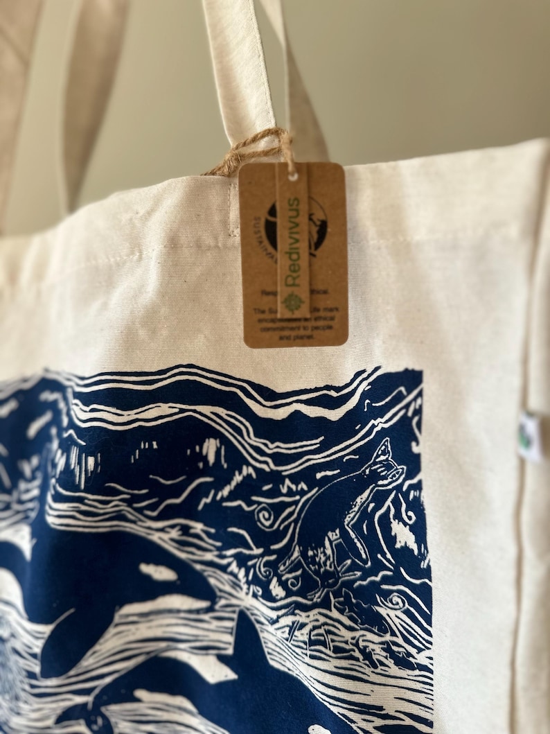 Puede incluir: Una bolsa de lona blanca con orcas y olas impresas en azul y blanco. La bolsa tiene una etiqueta de papel marr&oacute;n con la palabra "Redivvus" impresa en ella.