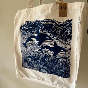 Puede incluir: Una bolsa de lona blanca con un estampado azul de dos orcas nadando en el oc&eacute;ano. El estampado incluye algas marinas, peces y otras formas de vida marina. La bolsa tiene una etiqueta marr&oacute;n con el texto "Redfinous" en ella.