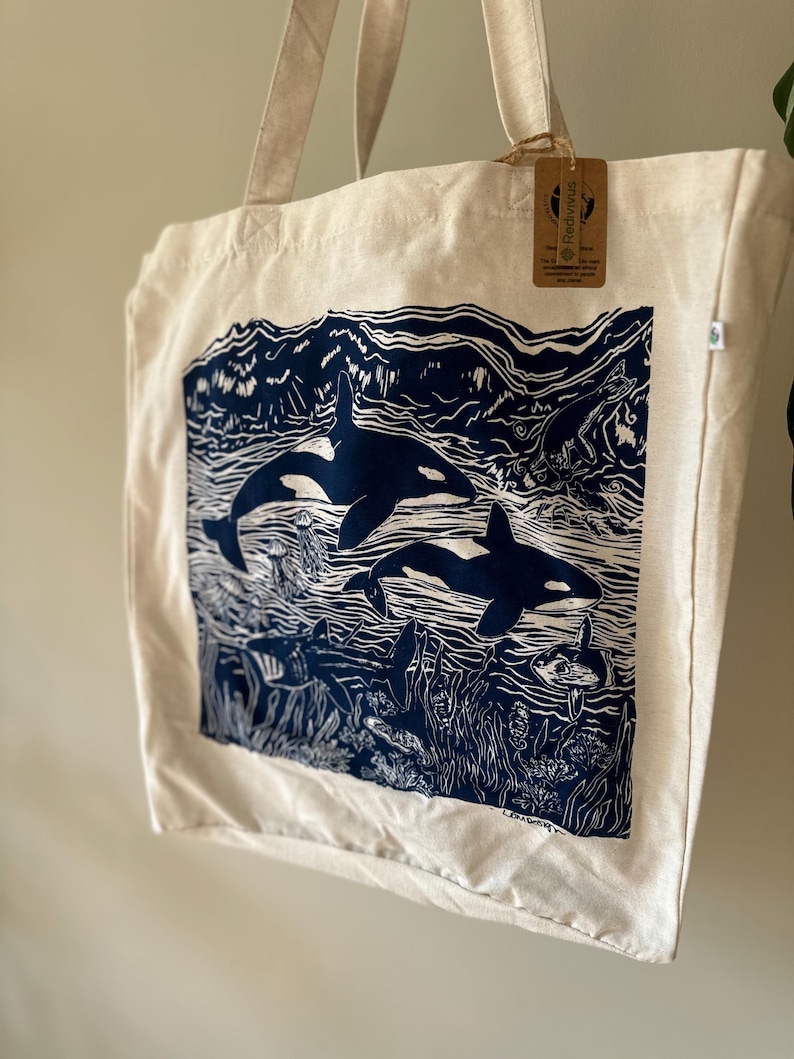 Puede incluir: Una bolsa de lona blanca con un estampado azul de dos orcas nadando en el oc&eacute;ano. El estampado incluye algas marinas, peces y otras formas de vida marina. La bolsa tiene una etiqueta marr&oacute;n con el texto "Redfinous" en ella.
