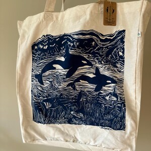 Puede incluir: Una bolsa de lona blanca con una ilustraci&oacute;n azul y blanca de orcas nadando en el oc&eacute;ano. La ilustraci&oacute;n incluye algas marinas, medusas y otras formas de vida marina. La bolsa tiene una etiqueta marr&oacute;n con el texto "FreddyWills" en ella.