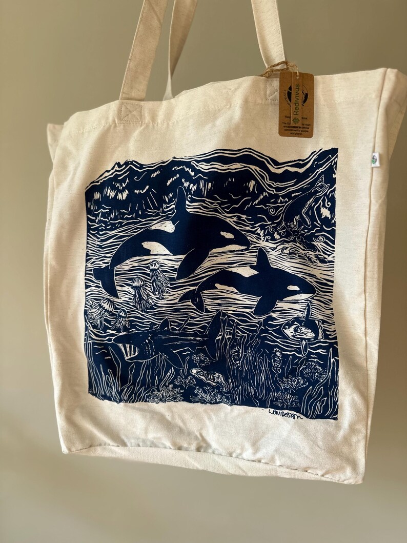 Puede incluir: Una bolsa de lona blanca con una ilustraci&oacute;n azul y blanca de orcas nadando en el oc&eacute;ano. La ilustraci&oacute;n incluye algas marinas, medusas y otras formas de vida marina. La bolsa tiene una etiqueta marr&oacute;n con el texto "FreddyWills" en ella.