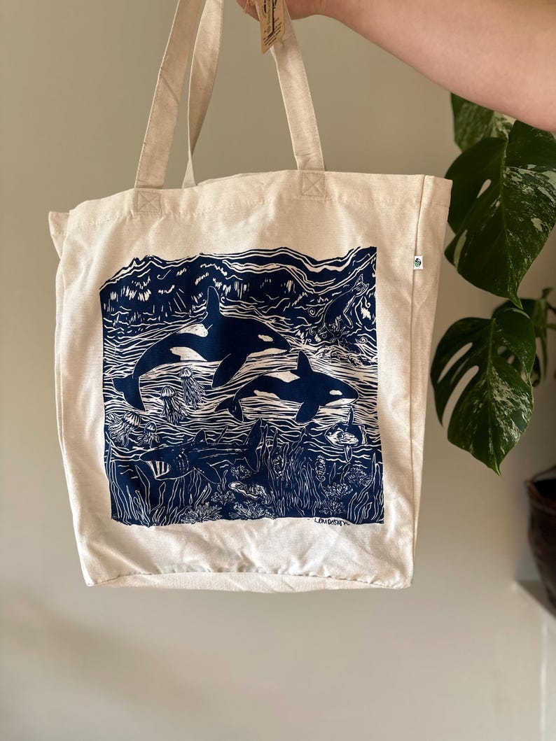 Puede incluir: Una bolsa de lona blanca con un estampado azul de orcas nadando en el oc&eacute;ano. El estampado incluye algas marinas, peces y otras formas de vida marina. La bolsa tiene un asa larga y est&aacute; hecha de algod&oacute;n natural.