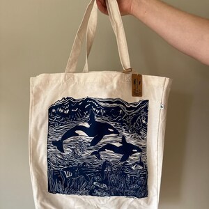 Puede incluir: Una bolsa de lona blanca con un estampado azul de dos orcas nadando en el oc&eacute;ano. El estampado tambi&eacute;n incluye algas marinas, peces y medusas. La bolsa tiene una etiqueta marr&oacute;n con el texto "Made in the USA" en ella.