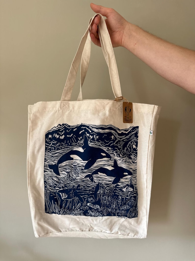 Puede incluir: Una bolsa de lona blanca con un estampado azul de dos orcas nadando en el oc&eacute;ano. El estampado tambi&eacute;n incluye algas marinas, peces y medusas. La bolsa tiene una etiqueta marr&oacute;n con el texto "Made in the USA" en ella.