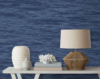 Puede incluir: Una mesa blanca con un jarrón de cerámica blanca, una escultura de coral blanco y una lámpara con una pantalla beige y una base tejida. La mesa está delante de una pared azul con un patrón ondulado.