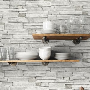 May include: A white brick wall with two wooden shelves holding various dishes and glasses. Below the shelves are ten glass jars filled with different types of grains and beans. A white bowl and a wooden cutting board are on the counter.